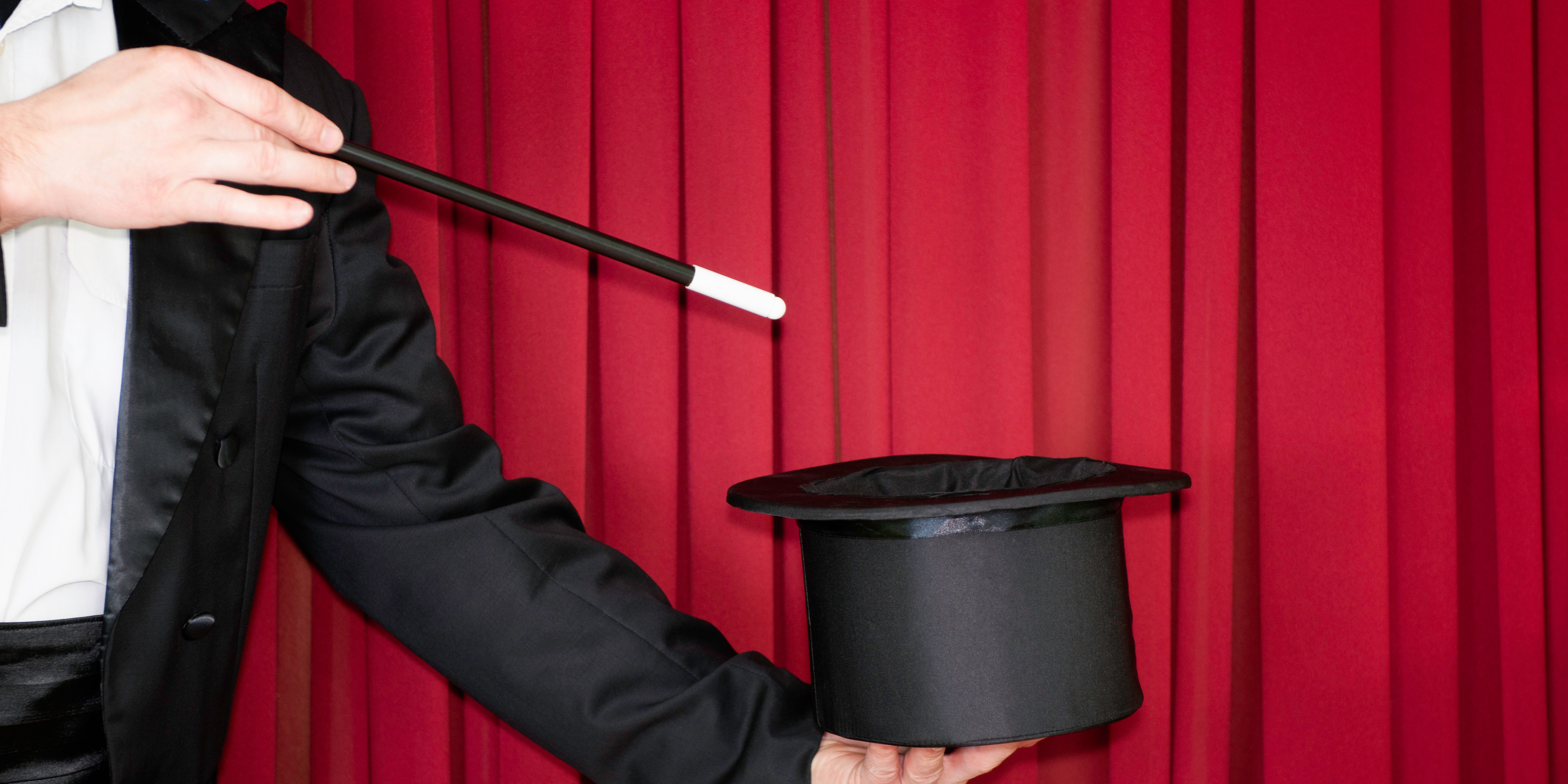 A magician's hand holding a wand performs a trick with a black top hat in front of a closed red curtain.