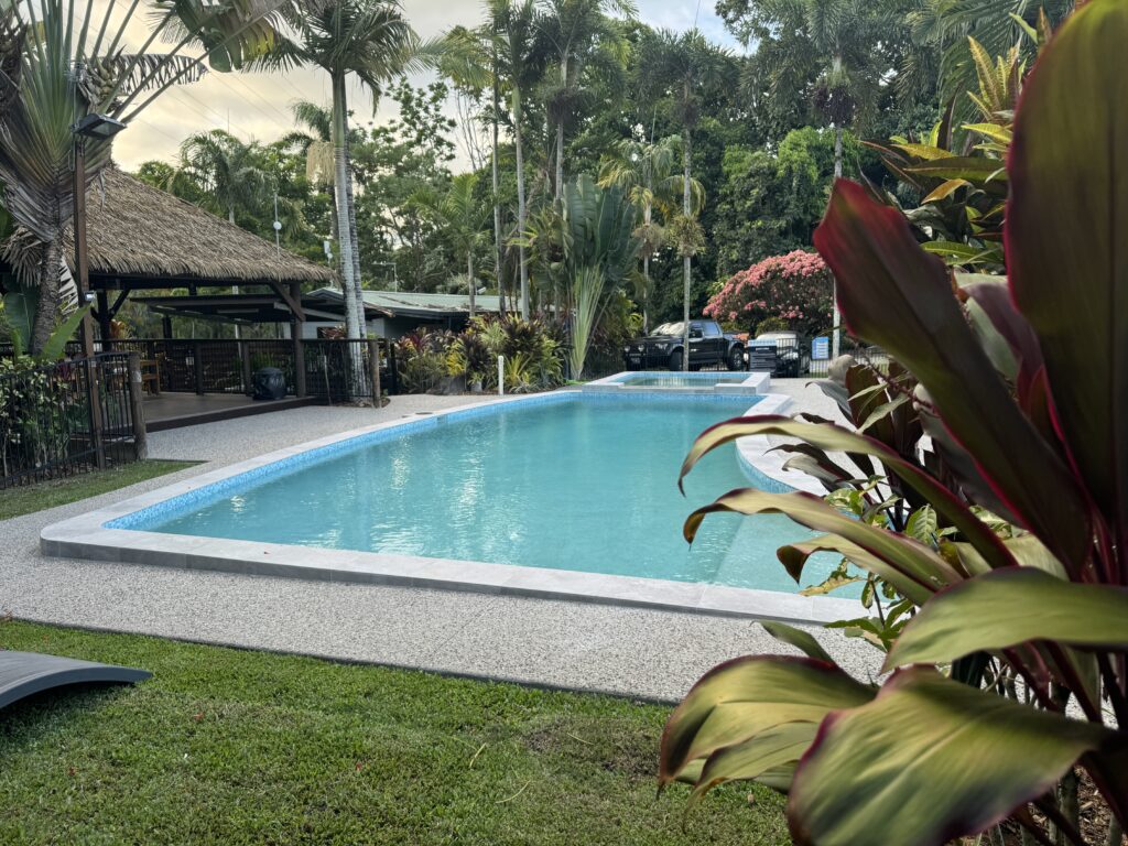 A serene outdoor pool surrounded by lush greenery and tall palm trees under a cloudy sky. A small thatched-roof structure stands to the left, with a parked car in the background. Vibrant plants frame the foreground.