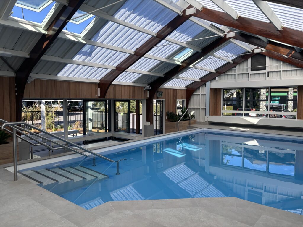 An indoor swimming pool with clear blue water is surrounded by a wooden deck, resembling the tranquility of a campsite retreat. The ceiling, made of glass panels, allows natural light to flood in. Metal handrails lead into the pool, and large windows offer picturesque views of trees outside.