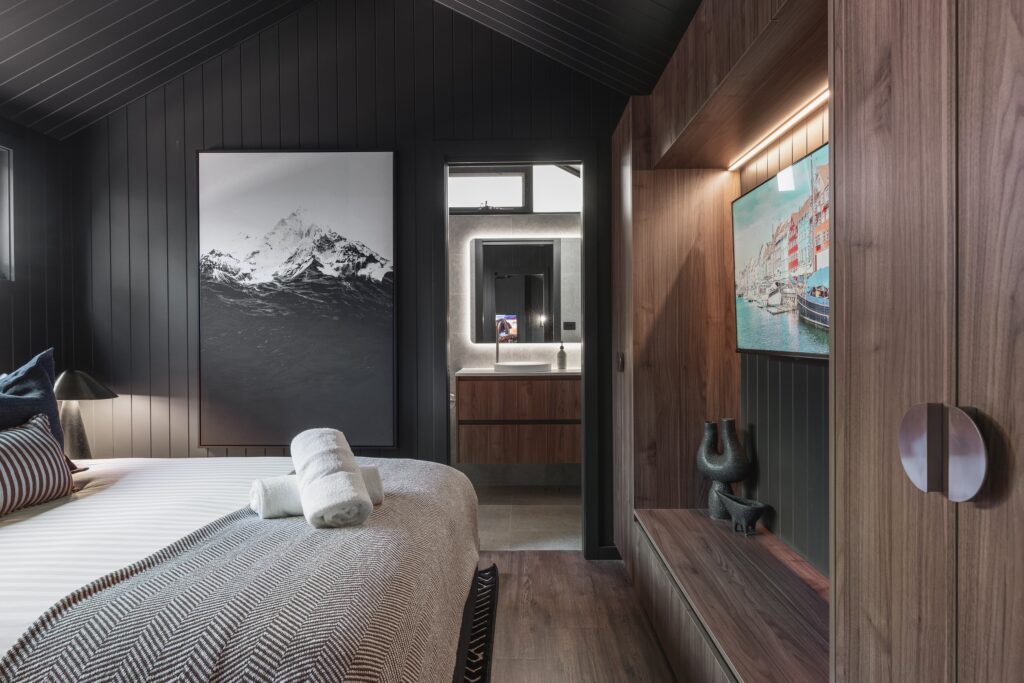 A cozy bedroom with dark wood paneling features a bed with striped pillows and a herringbone blanket. A large mountain photo is on the wall. The doorway reveals a bathroom with a mirror reflecting soft light. A painting of a harbor scene is on the right.