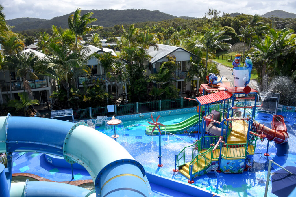 A colorful water park features slides, splash buckets, and climbing structures. Surrounded by tropical trees and hills, it's a perfect camping getaway. Nearby two-story buildings complement the lush, adventurous environment where you'll need all your gear for a day of fun.