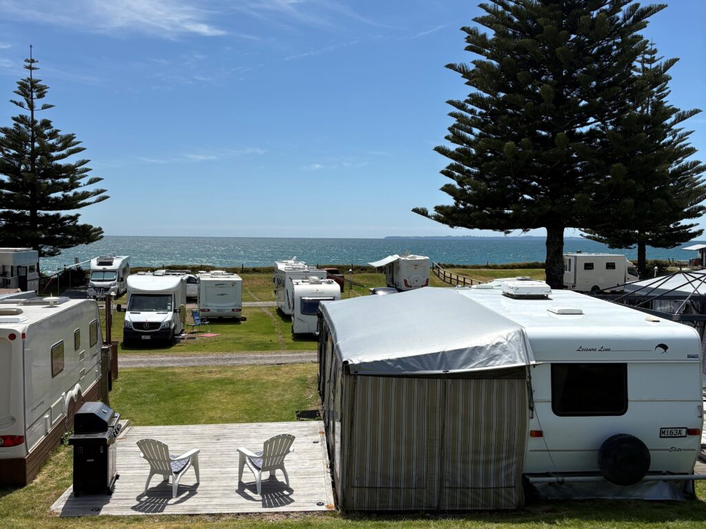 A scenic view of a coastal RV park with several campers and trailers. Two empty chairs are on a wooden deck in the foreground, facing the ocean. Tall pine trees dot the grassy area, and the sky is clear and blue.