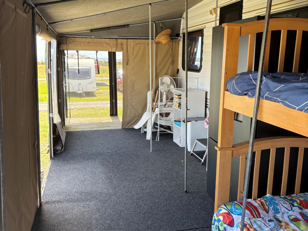 The image shows the interior of a camper with a carpeted floor. It has bunk beds on the right, a small table with chairs, and camping gear. The area is enclosed with fabric walls, and there's an open view to the outside.