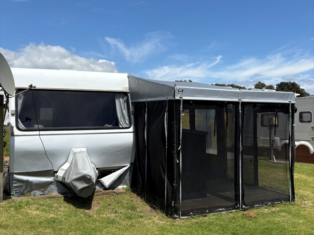 A camper van with a black mesh extension attached, parked on a grassy field. A satellite dish is visible on the left, under a clear blue sky with a few clouds.