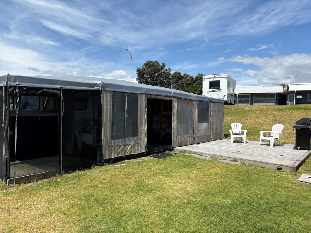 A large tent with open sides is set up on wooden decking in a grassy area. Two white outdoor chairs face a barbecue grill under a clear blue sky. In the background, there is a white RV parked.