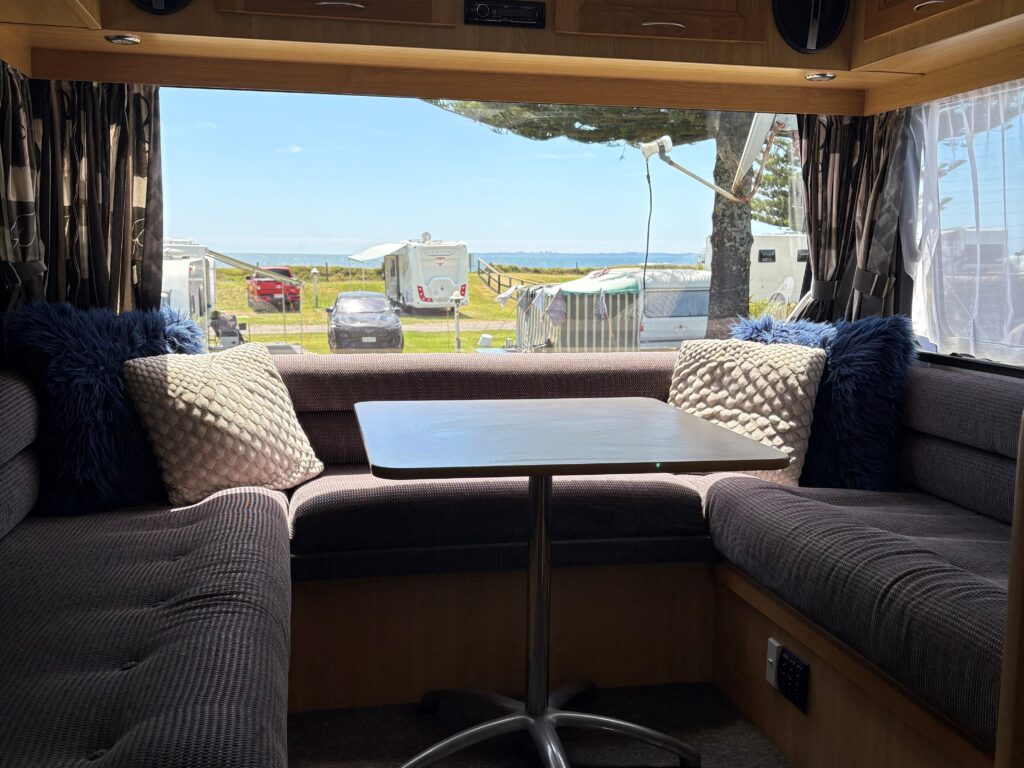 An interior view of a camper van shows a cozy seating area with a table and cushioned benches. Two windows offer a scenic view of parked trailers, green grass, and a clear blue sky outside.