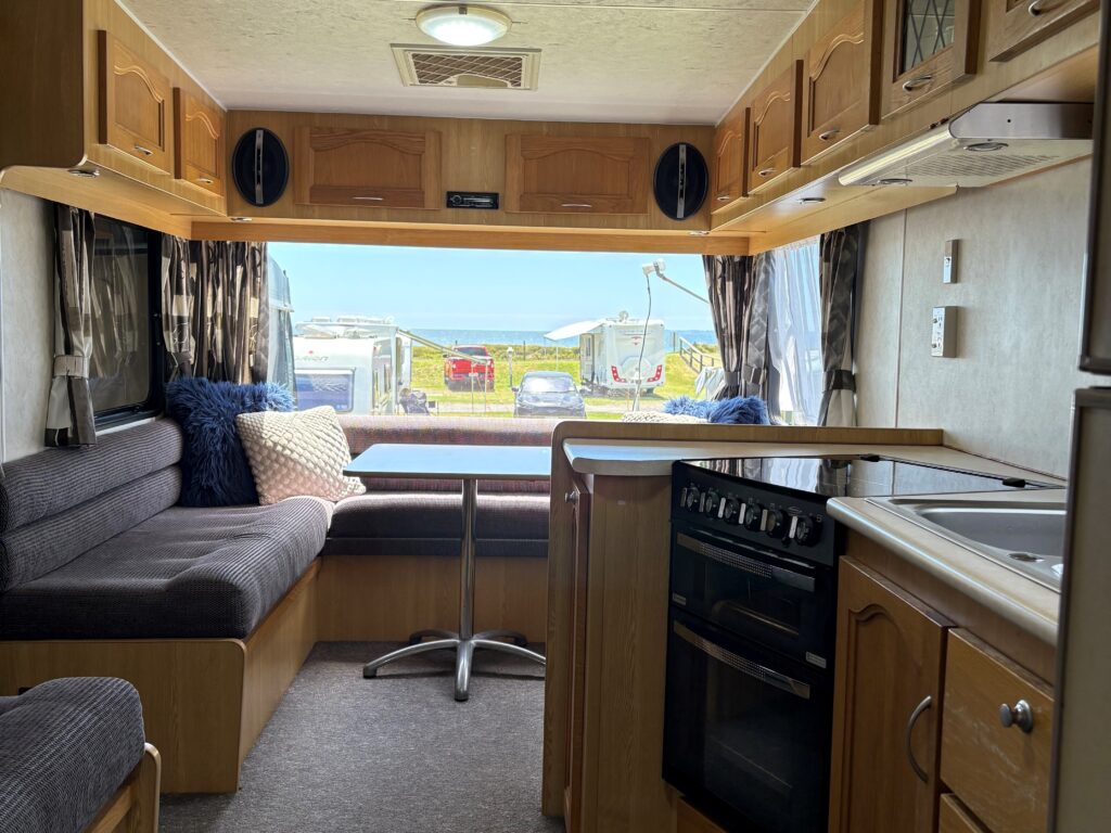 Interior of a cozy caravan featuring a kitchen with wooden cabinets, a stove, and a sink. A seating area with gray cushions and pillows is on the left. The window at the end reveals a view of a grassy area and the ocean in the distance.