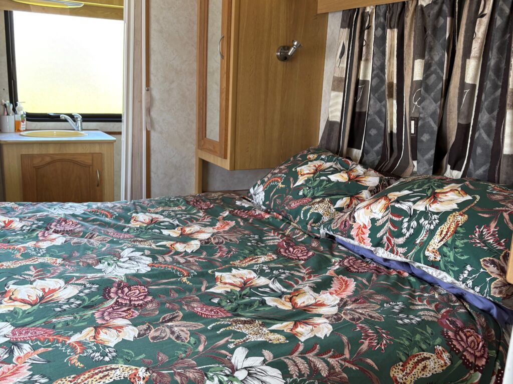 A cozy bedroom inside a camper van features a floral-patterned bedspread, wooden cabinets, and a window with patterned curtains. The compact space also includes a glimpse of a small kitchenette.