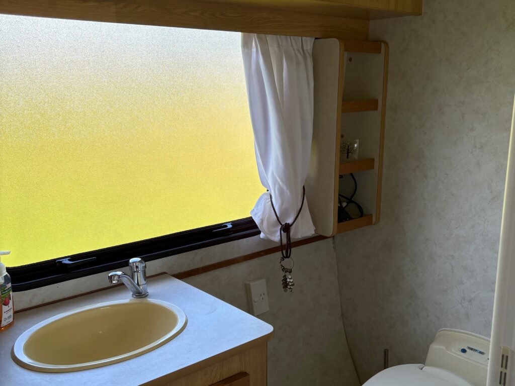 Interior of a compact RV bathroom featuring a white toilet and a beige sink with a single faucet on a wooden countertop. A frosted window with a white curtain is above the sink, providing natural light. Cabinets and a small shelf are mounted on the walls.