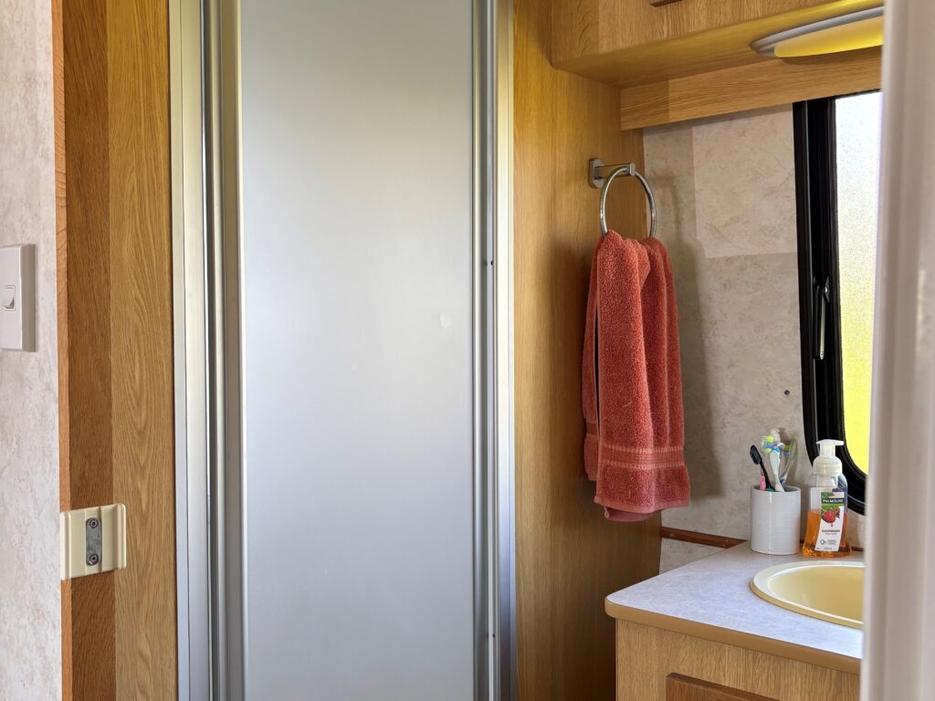 Interior of a small bathroom with wooden cabinets. A glass shower door is on the left, a pink towel hangs on a ring, and toiletries are arranged near a round sink with a faucet. A window is above the counter.