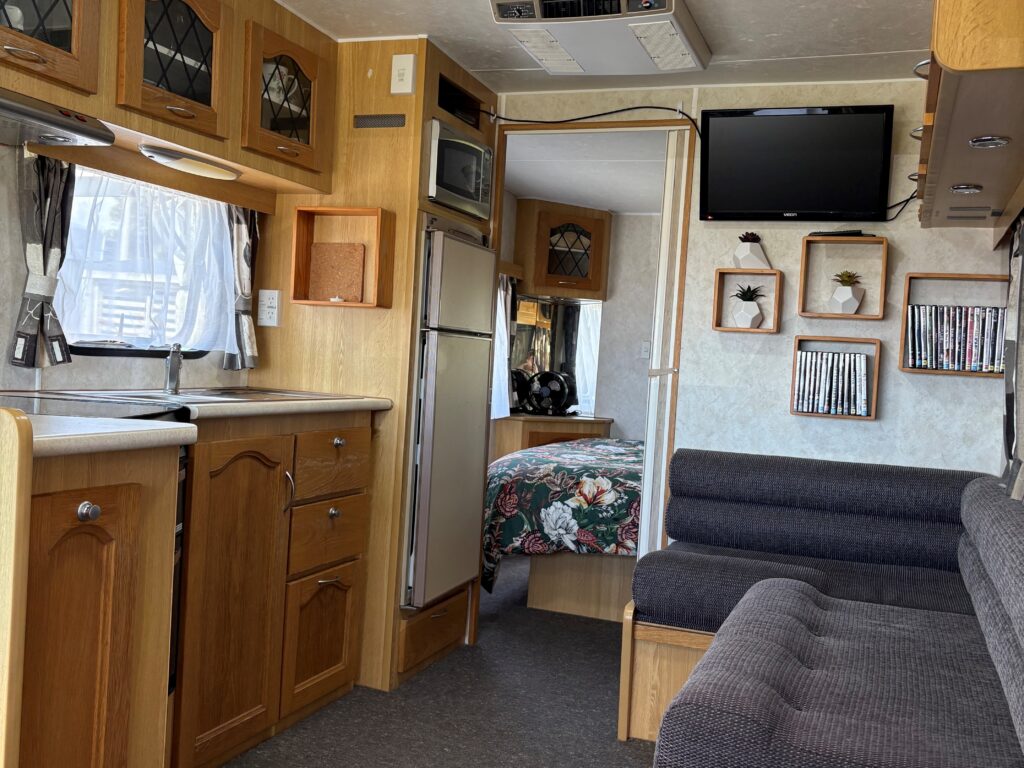 Interior of a cozy RV with wooden cabinets, a small kitchenette, and a seating area. A bed is visible in the background. The walls have shelves with decorations and a mounted TV. Natural light enters through curtained windows.