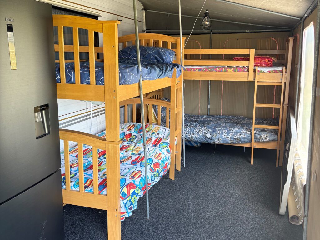 Interior of a tent or cabin with two wooden bunk beds, featuring colorful bedding. A black refrigerator is positioned on the left side, and the floor is carpeted in dark gray. The setting suggests a camp or temporary accommodation.