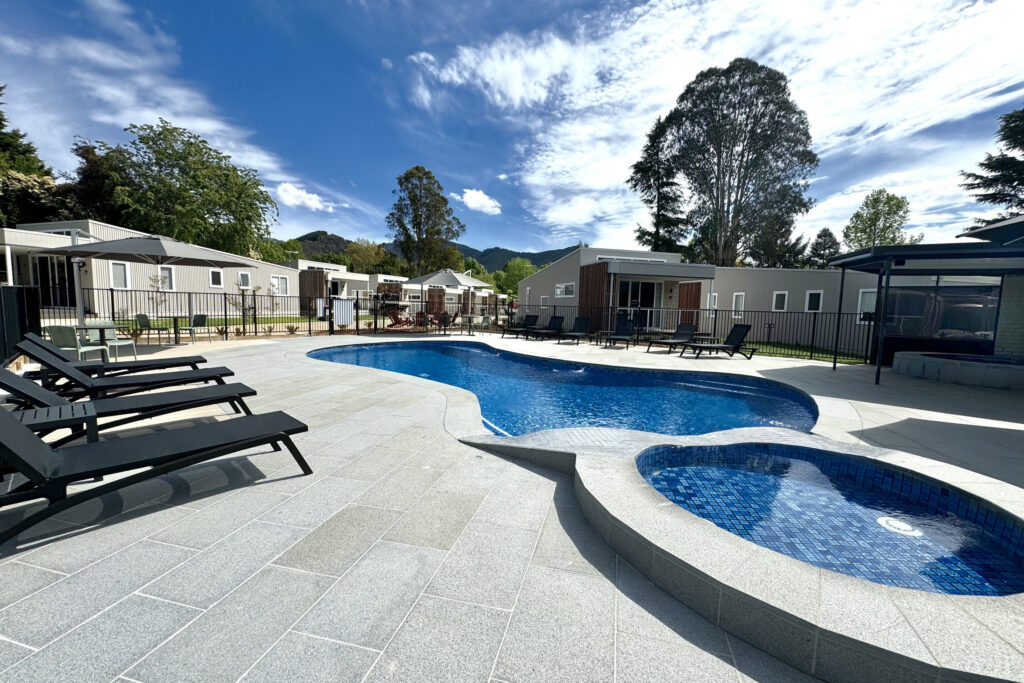 A modern outdoor pool area features a uniquely shaped swimming pool and an adjacent round hot tub, perfectly equipped for gear stowaway. Surrounding are several lounge chairs, ideal for those who prefer comfort after a day of camping. Small buildings and tall trees stand under a partly cloudy sky.