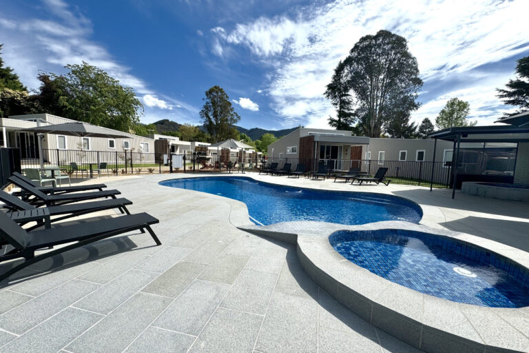 Outdoor pool area with a curvy blue swimming pool and a smaller circular spa at Retreat on Delany. Surrounded by lounge chairs and modern buildings. Tall trees and a mountain are visible in the background under a partly cloudy sky.