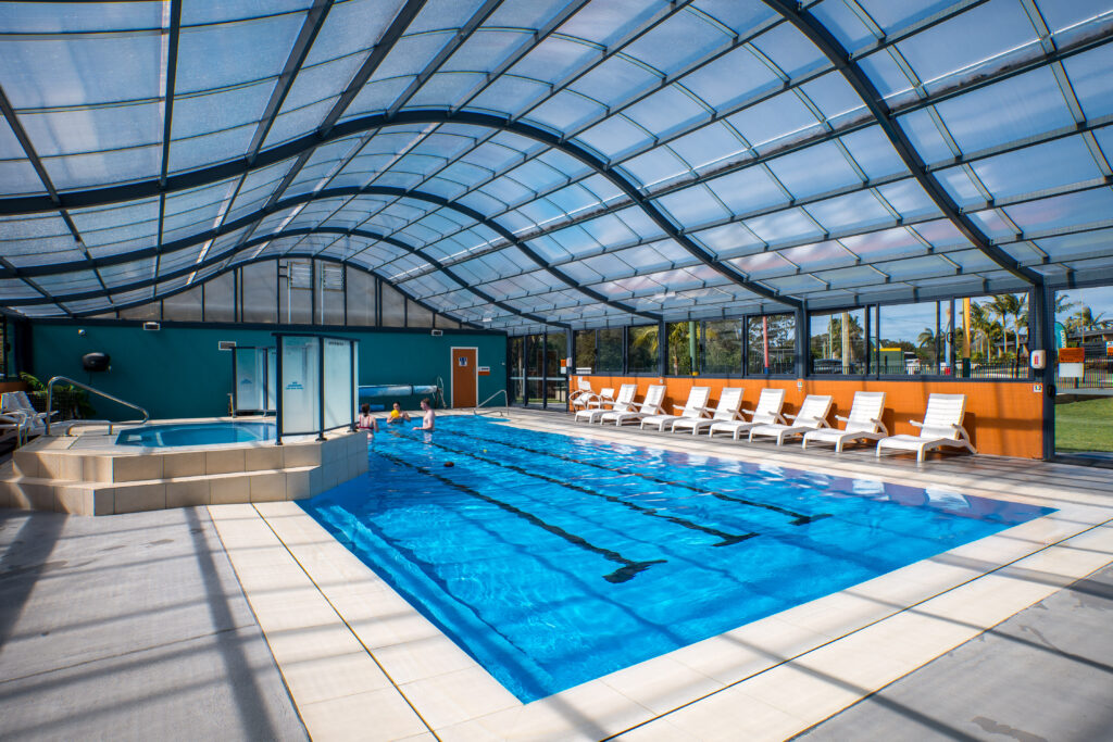 The indoor swimming pool, with its curved glass roof and blue tiles, features designated swim lanes. Lounge chairs line the side near the expansive windows, offering views of green grass and trees outside—a perfect spot for relaxation before your next camping adventure requiring sturdy gear.