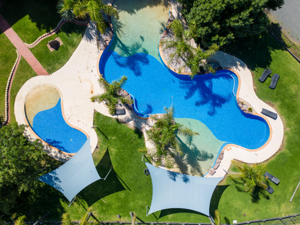 Aerial view of a uniquely shaped swimming pool surrounded by greenery. The pool includes a small circular section, with lounge chairs and trees around it. Two white sunshades resemble camping gear, positioned over a grassy area to provide shade.