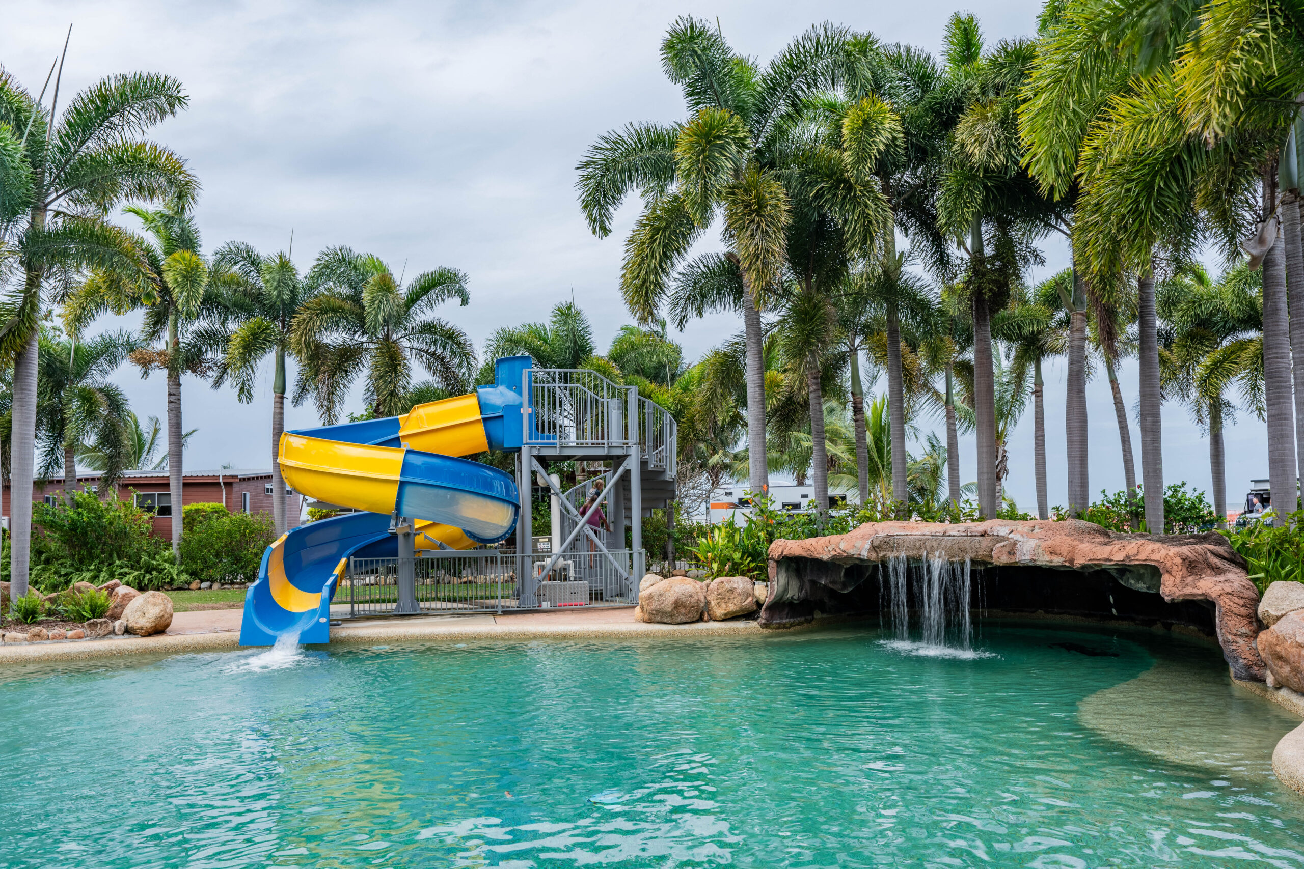 A tropical swimming pool scene features a spiral blue and yellow waterslide leading into the pool. Palm trees surround the area like nature's own camping gear, with a waterfall flowing from a rock formation on the right. The sky is overcast.