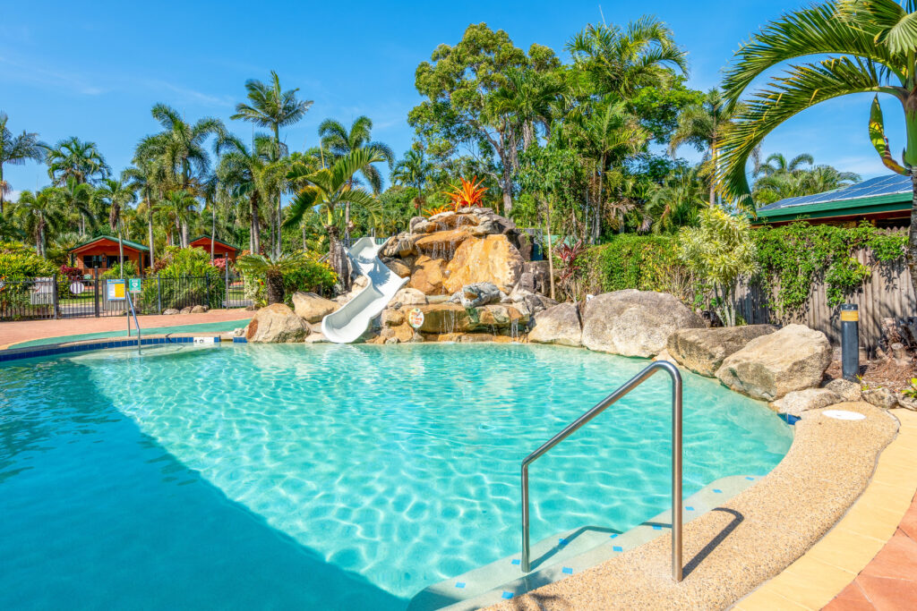 A tropical swimming pool with clear blue water features a small water slide surrounded by rocks and lush palm trees, perfect for a camping experience. A metal handrail leads into the pool, and two wooden cabins stocked with gear are visible in the background under a bright blue sky.