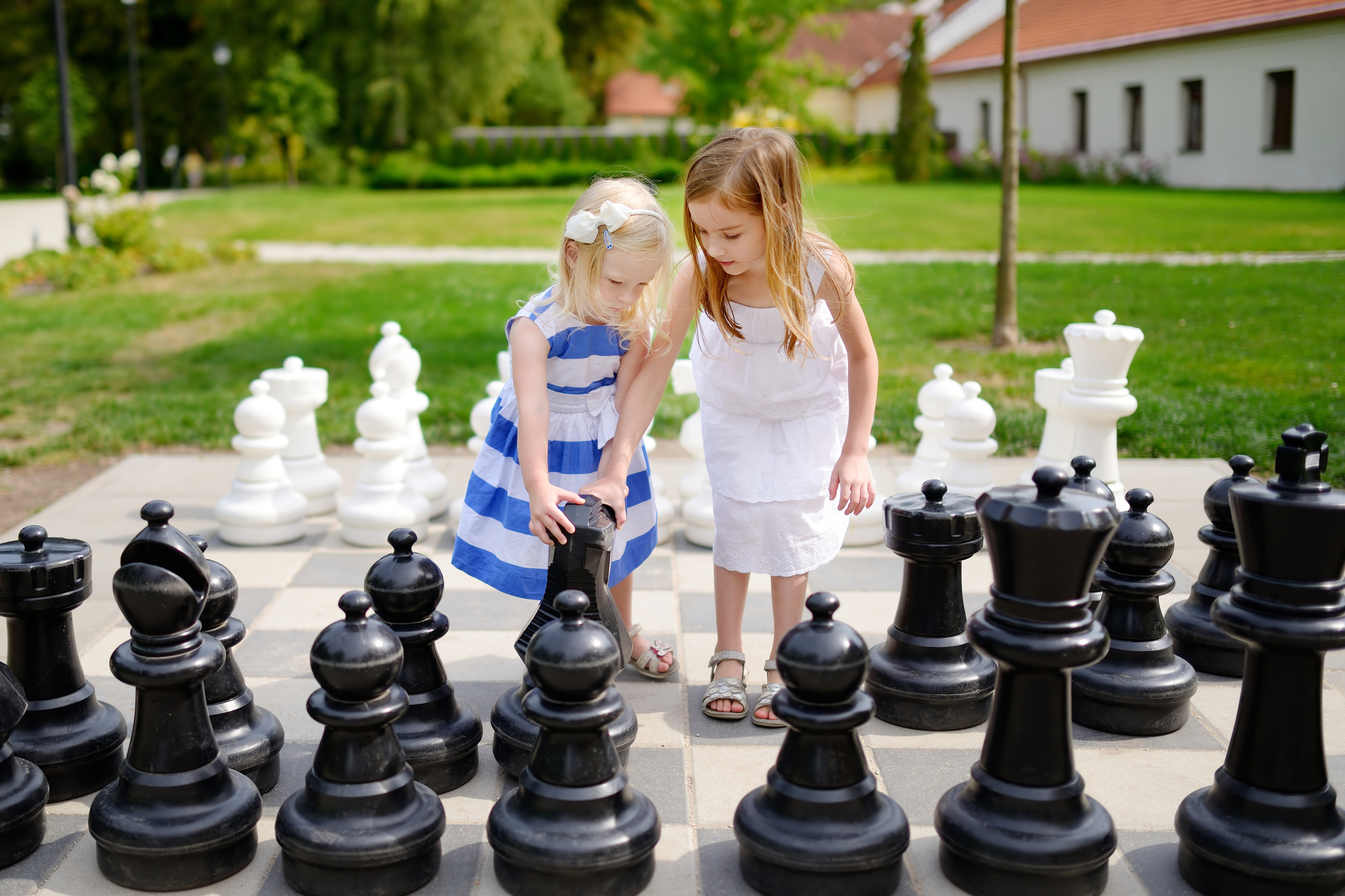 Two young girls, one in a blue striped dress and the other in a white dress, are playing with large black and white chess pieces in an outdoor garden setting.