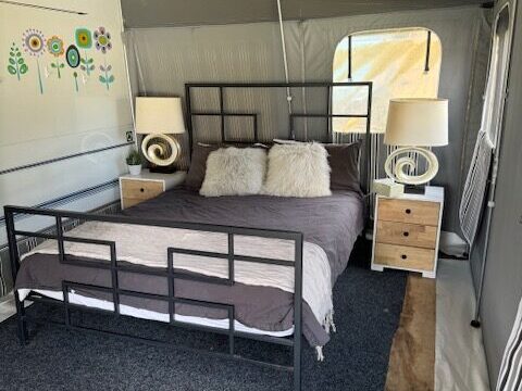 A cozy tent interior with a neatly made bed featuring a metal frame and decorative pillows. Two lamps sit on wooden nightstands beside the bed. The tent walls display whimsical flower drawings, adding a touch of color. A small window lets in light.