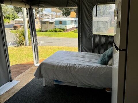 Interior of a tent with a bed, pillows, and a refrigerator. The tent has open sides revealing a grassy area and nearby structures under a cloudy sky.