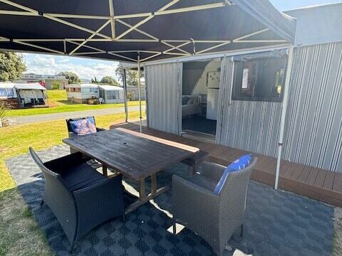 Outdoor seating area with a wooden table and wicker chairs under a large canopy. The setup is on a patterned mat beside a building, with a grassy lawn and clear sky in the background.