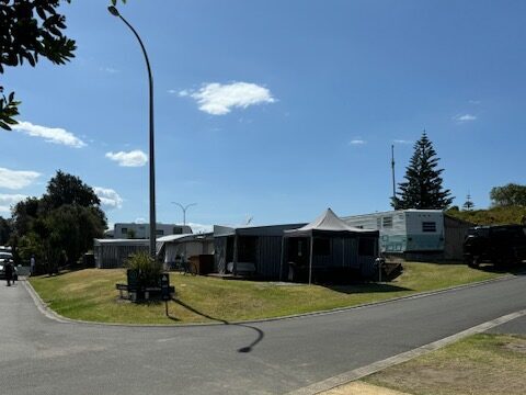 A sunny day in a suburban neighborhood with a view of a small park area. There are several caravans and trailers parked on the grass, with a few trees and blue sky with scattered clouds in the background. A street runs alongside the park.