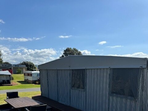 A caravan setup with a metal awning and outdoor patio features a wooden table and black chairs on a paved surface. It's situated on a grassy area under a clear blue sky. Other caravans and trees are visible in the background.