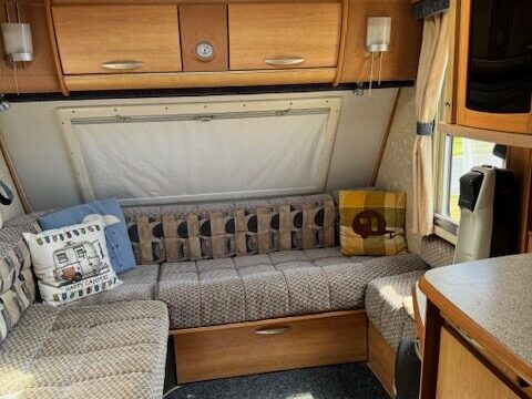 Cozy caravan interior with a skylight, featuring an L-shaped sofa with patterned cushions, wooden cabinets, and a compact kitchenette area. Sunlight streams in, highlighting the warm wood tones and comfortable layout.