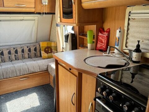 Interior of a cozy caravan kitchen with wood cabinets, a small sink, and a stove. A cushioned bench sits by the window, complete with patterned pillows and a folded blanket. Natural light brightens the space through a skylight.