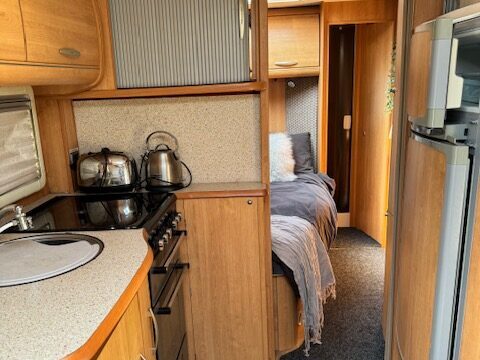 Interior of a camper van featuring a compact kitchen with a countertop, stove, and kettle. Beyond the kitchen is a sleeping area with a neatly made bed, offering a cozy and efficient living space.