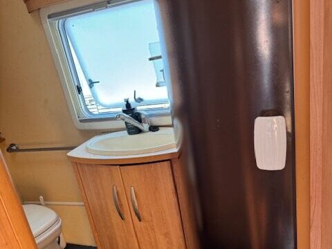 A compact bathroom in an RV featuring a white toilet, a small wooden vanity with a sink, and a mirror. There's a window providing natural light and a black cylindrical shower unit next to the sink. The floor is covered with dark carpeting.