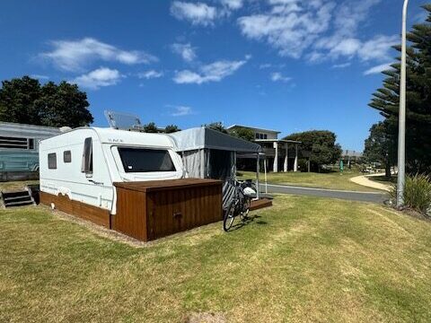 A white caravan with a wooden base is parked on grass under a blue sky with some clouds. A bicycle is leaning against it. Nearby are other caravans and trees. A pathway and streetlamp are in the background.