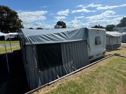 A caravan with an attached awning is parked on a grassy area under a partly cloudy sky. Nearby, additional caravans and trees are visible, suggesting a campsite setting.