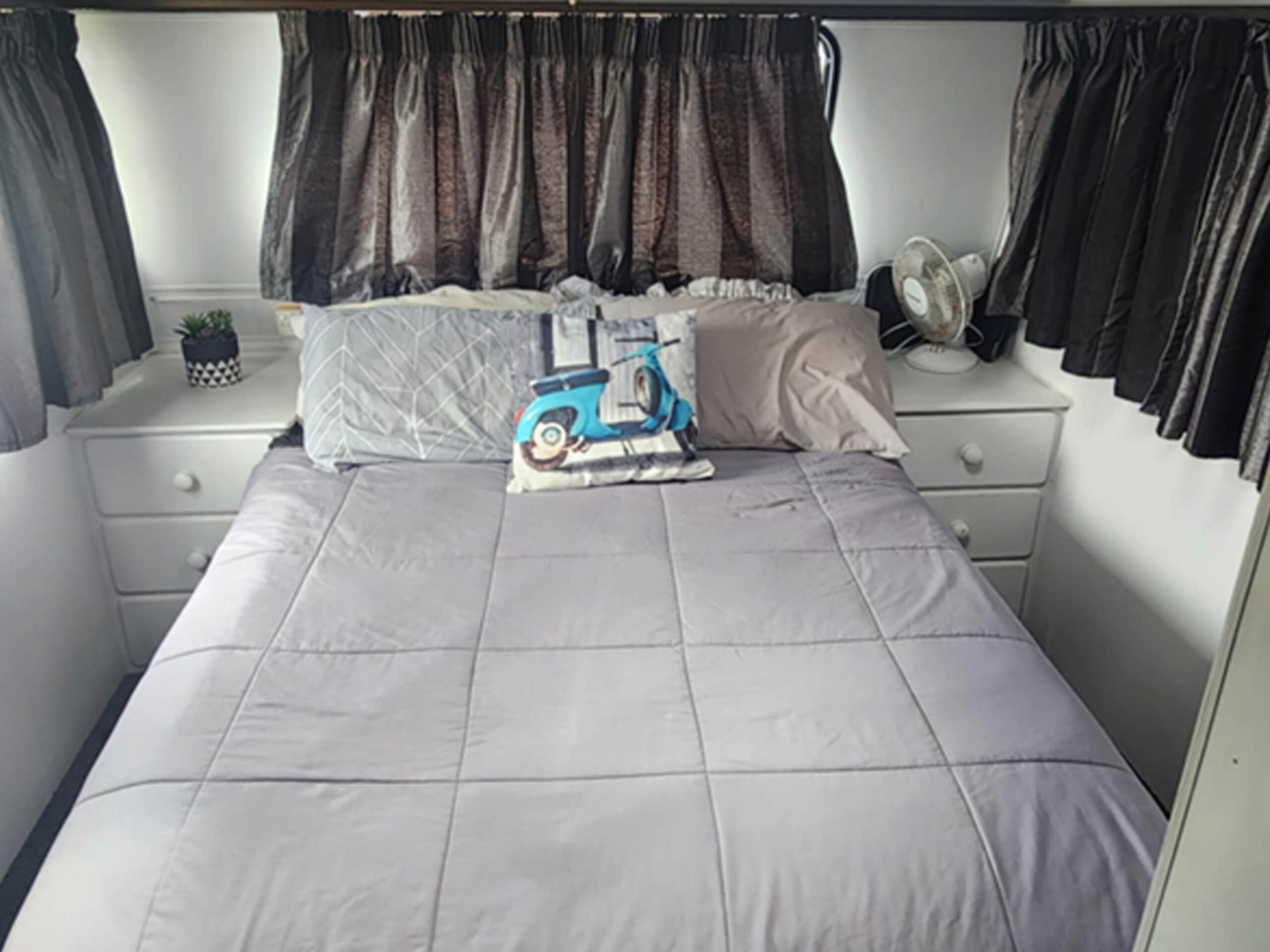 A cozy bedroom features a neatly made bed with a light gray comforter, several pillows including one with a scooter design, white bedside tables, a small fan, and a potted plant. Dark curtains hang over the windows, giving a warm and inviting feel.