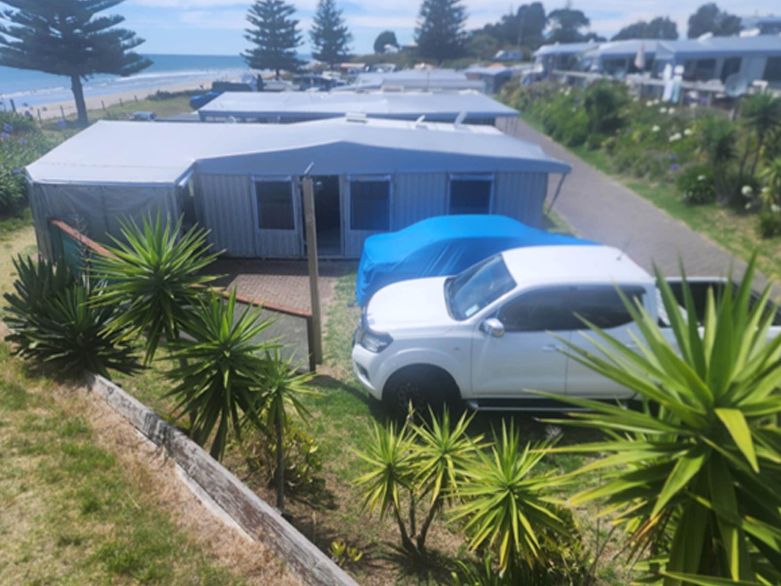 Coastal campsite with several caravans and parked vehicles. A white SUV is covered with a blue tarp. The area is lined with greenery and trees, and the ocean is visible in the background, providing a scenic view.