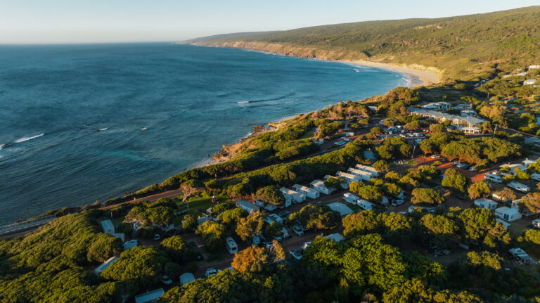 Aerial view of a coastal landscape with lush green hills and a winding beachline. The scene showcases camping gear at a campsite with caravans and small buildings nestled among dense trees, adjacent to the turquoise ocean waves.