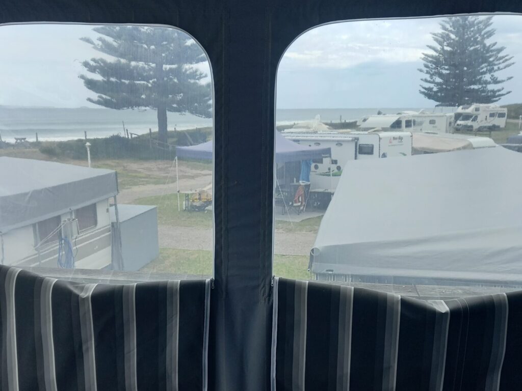 View through a window showing a campground with several caravans and tents. Two tall pine trees are visible in the background near the ocean under a cloudy sky. The window has striped curtains partly covering the lower half.