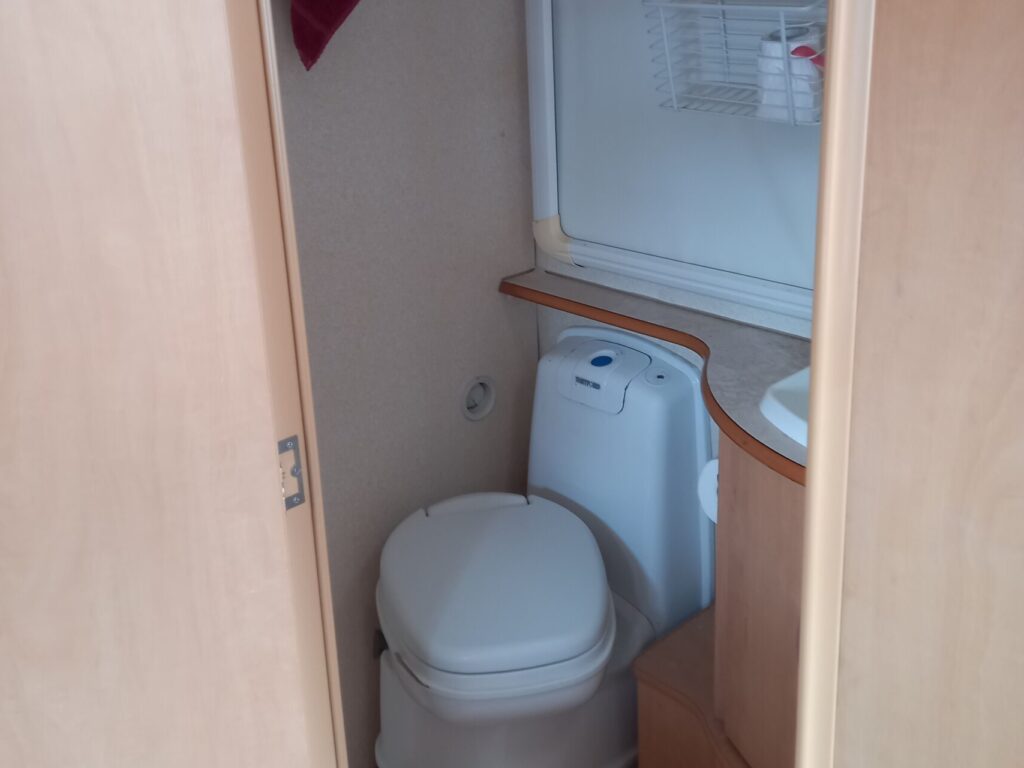 A small bathroom with a toilet, sink, and open shelving. The shelves hold various toiletries, and a red towel hangs from a hook on the wall. The floor is light wood, and the door is slightly ajar.