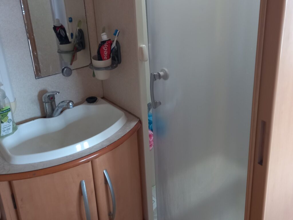 A small bathroom with a frosted glass shower enclosure and a wooden sink cabinet. The sink has a faucet, a mirror above, and toiletries. A liquid soap dispenser is visible on the counter.