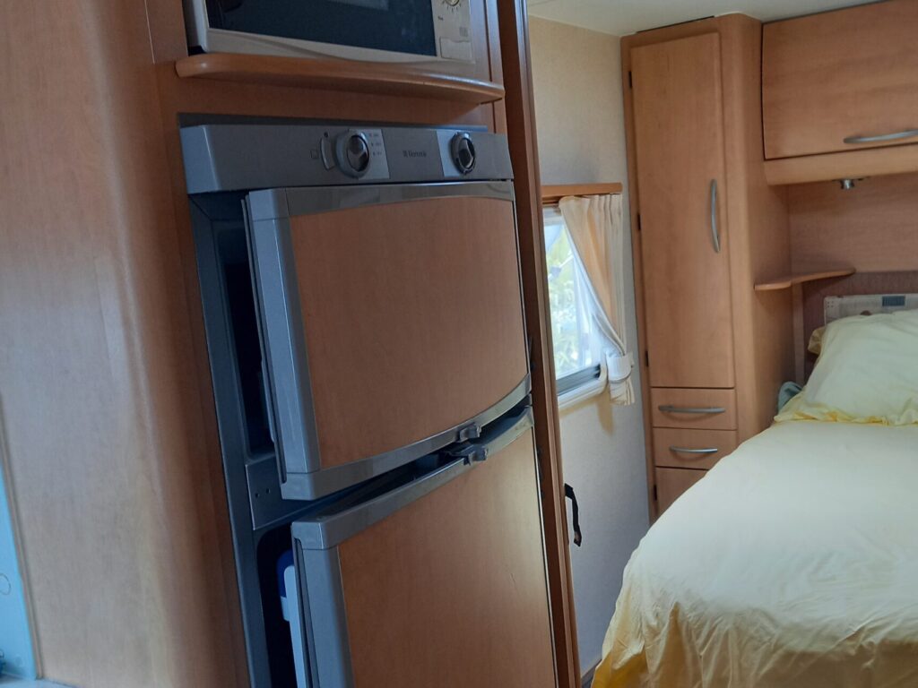 Interior of a camper, showing a compact kitchen with a fridge, microwave, and stove. A cozy bed with yellow bedding is visible in the background, under a skylight, with wood-paneled storage cabinets surrounding the area.