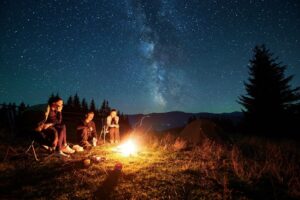 Three campers sit around a campfire at night, enjoying the warmth and stargazing in the clear sky above.